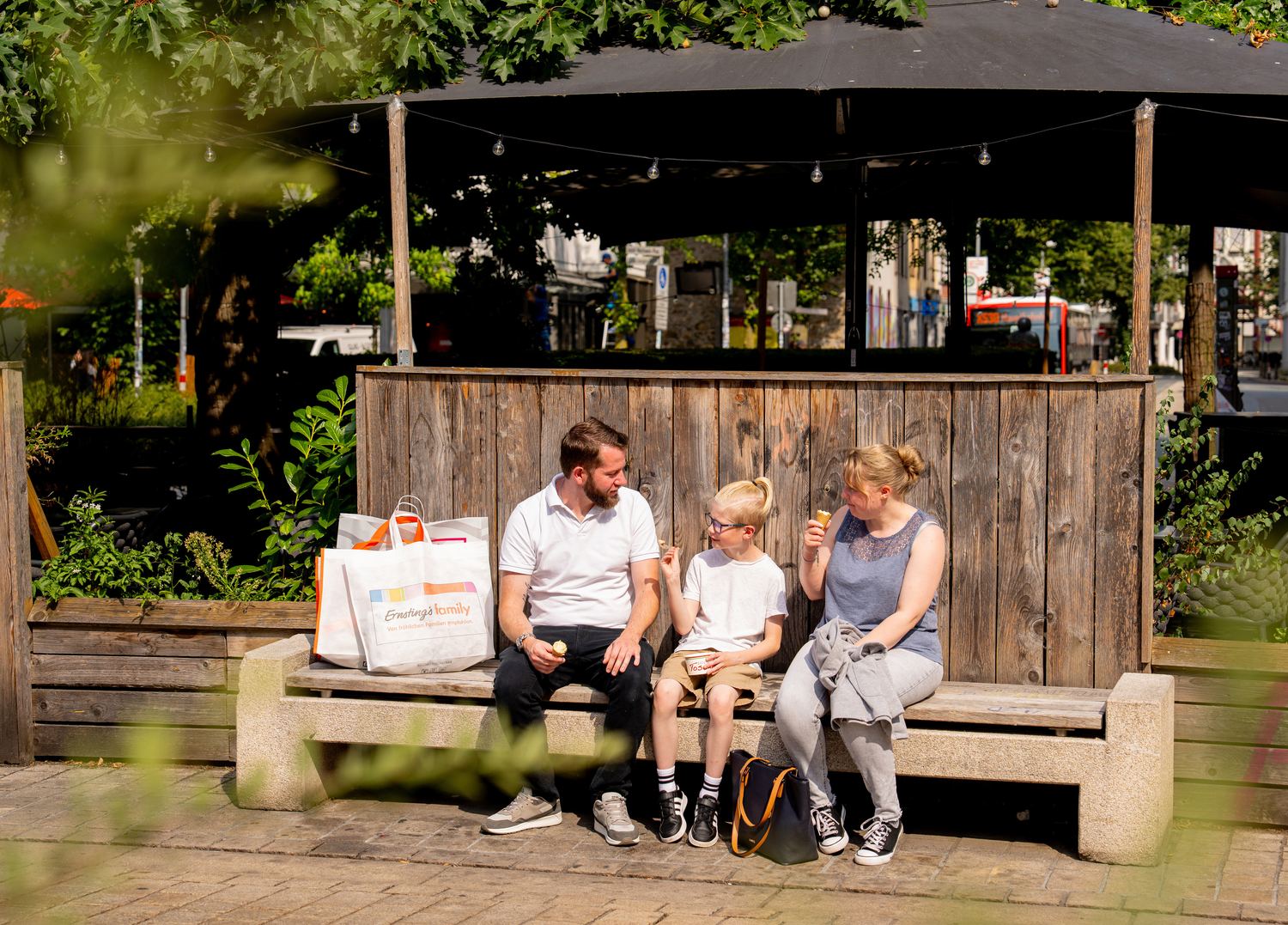 Die Familie sitzt mit Einkaufstaschen und Eis auf einer Holzbank unter einem Pavillon.
